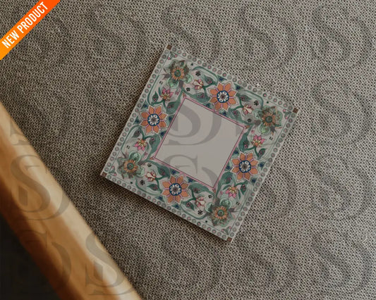 Square wedding card with Mughal-inspired floral border placed on a textured beige fabric surface in a flatlay arrangement.