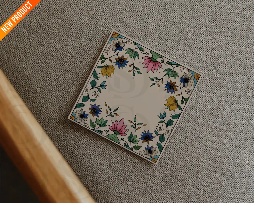 Square wedding card with a Mughal floral frame placed on a textured beige fabric in a flatlay presentation.