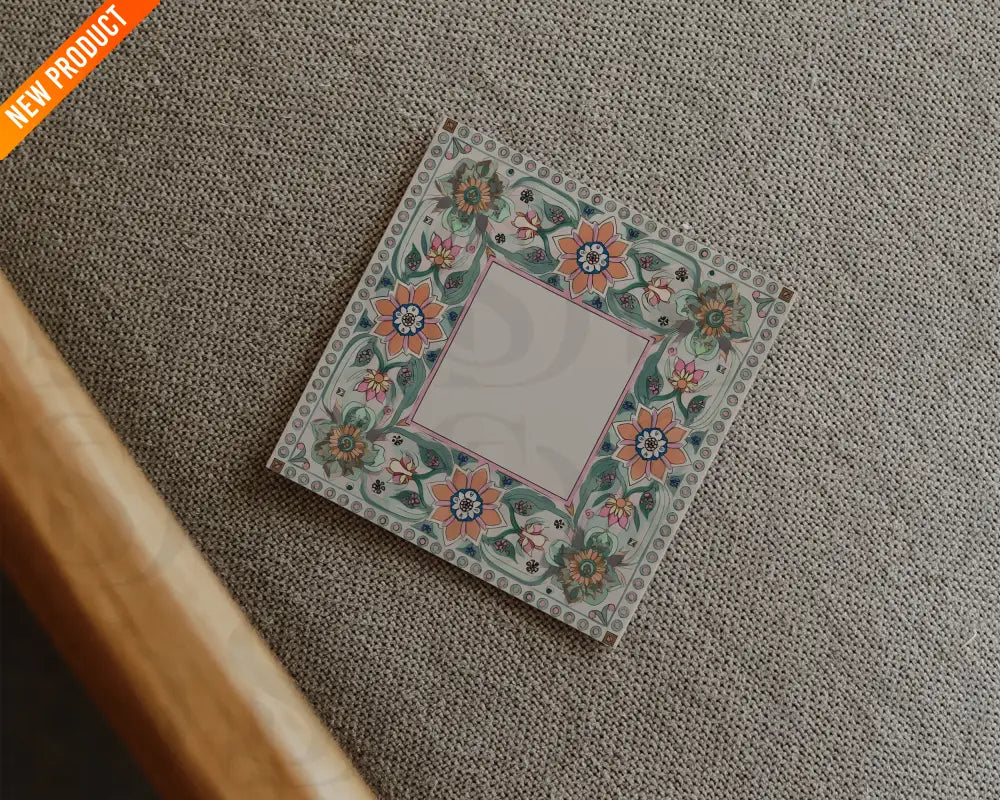 Square wedding card with Mughal-inspired floral border placed on a textured beige fabric surface in a flatlay arrangement.
