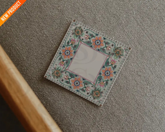 Square wedding card with Mughal-inspired floral border placed on a textured beige fabric surface in a flatlay arrangement.