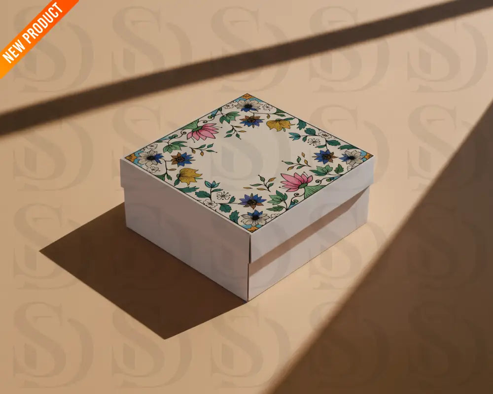 Square gift box with Mughal-inspired floral frame design on the lid, featuring lotus and botanical motifs, photographed on a beige surface with shadows.