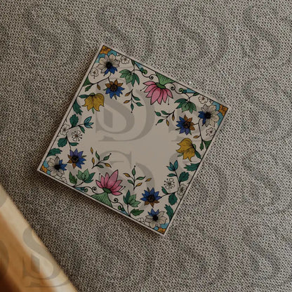 Square wedding card with a Mughal floral frame placed on a textured beige fabric in a flatlay presentation.