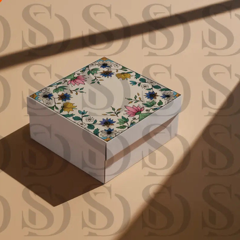 Square gift box with Mughal-inspired floral frame design on the lid, featuring lotus and botanical motifs, photographed on a beige surface with shadows.