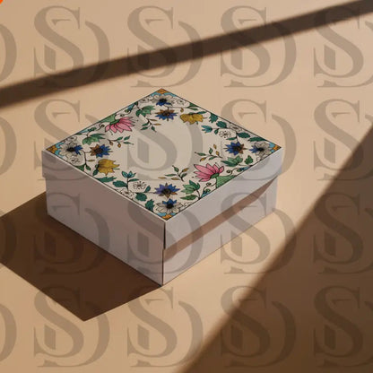 Square gift box with Mughal-inspired floral frame design on the lid, featuring lotus and botanical motifs, photographed on a beige surface with shadows.